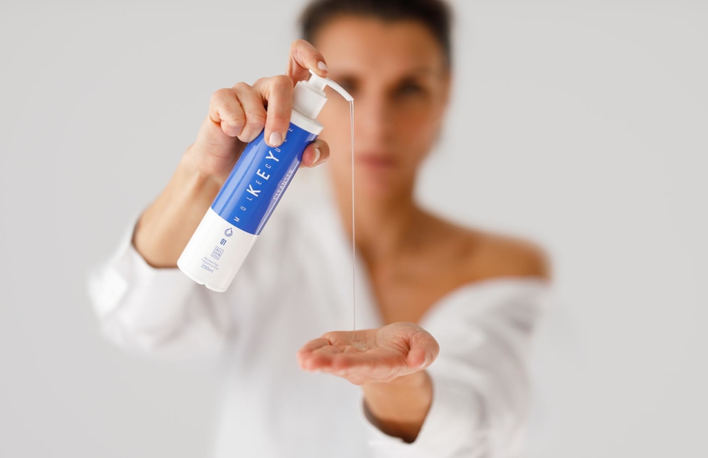 Studio shot of a young woman in a white shirt, applying a skincare lotion to the palm of her hand. Focus on the product.
